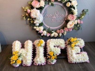 Yellow Wife Letters - Wife letter tribute, arranged with a Pink ribbon edge, white based flowers and yellow letter sprays. These can be arranged in your own choice of colours.